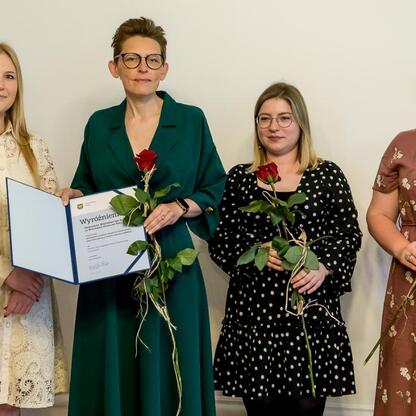 Czterech kobiet w eleganckiej odzieży, trzymające certyfikaty i czerwone róże, uczestniczą w uroczystości wręczenia Nagród Marszałka Województwa Śląskiego za wydarzenie muzealne 2024. (wygenerowano przez AI)