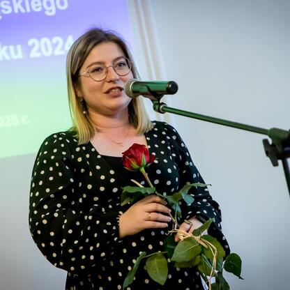 Podczas uroczystości wręczenia Nagród Marszałka Województwa Śląskiego za Wydarzenie Muzealne roku 2024, uczestniczka przemawia na podium, trzymając bukiet czerwonych róż. (wygenerowano przez AI)