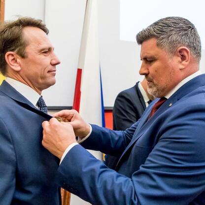 Dwóch mężczyzn w strojach formalnych, jednego nagradzającego drugiego medalem, w tle flagi i tablica, w ramach Tygodnia Kultury Beskidzkiej. (wygenerowano przez AI)