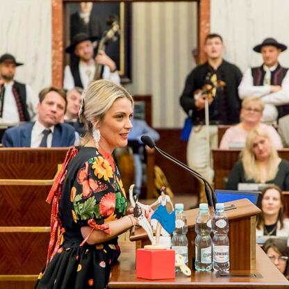 W czasie XV sesji Sejmiku Województwa Śląskiego, kobieta w kwiatowym sukni prezentuje figurkę i nożyczki, podczas uroczystości kulturalnej Tydzień Kultury Beskidzkiej. (wygenerowano przez AI)