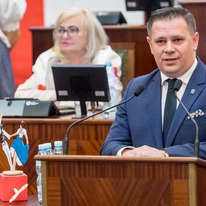 W Sejmiku Województwa Śląskiego, podczas Tygodnia Kultury Beskidzkiej, mężczyzna w garniturze przemawia do mikrofonu, za nim siedzi kobieta w białym płaszczu. (wygenerowano przez AI)