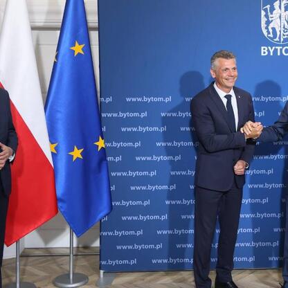 Trzech mężczyzn w strojach formalnych trzyma się za ręce przed logo i stroną internetową Bytom, Polska, z flagami UE i Polski. (wygenerowano przez AI)
