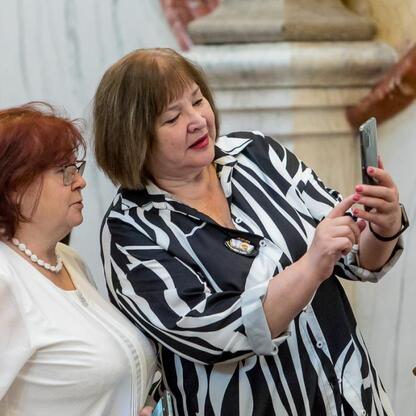 Dwie kobiety w strojach formalnych robią selfie w sali konferencyjnej, podczas 50-lecia Uniwersytetów Trzeciego Wieku w Polsce. (wygenerowano przez AI)