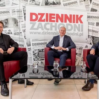 Trzech mężczyzn dyskutuje w modernistycznym biurze Dziennika Zachodniego, omawiając przyszłość edukacji i nauki w Śląsku. (wygenerowano przez AI)