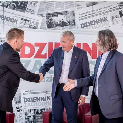 Trzech mężczyzn w strojach formalnych trzyma się za ręce przed tłem z tytułami 
