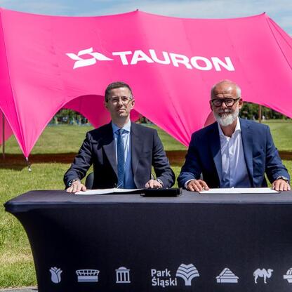 Dwa mężczyźni w strojach formalnych siedzą przy stole z logo TAURON Park Śląski, pod dużym różowym namiotem. (wygenerowano przez AI)