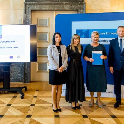 Grupa czterech osób prezentuje projekt finansowany z Europejskiego Funduszu Rozwoju Regionalnego dla Gminy Ogrodzieniec, o wartości 1,628,076.19 PLN. (wygenerowano przez AI)