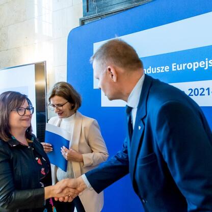 Dwa osoby w strojach formalnych składają gratulacje na tle transparentu 