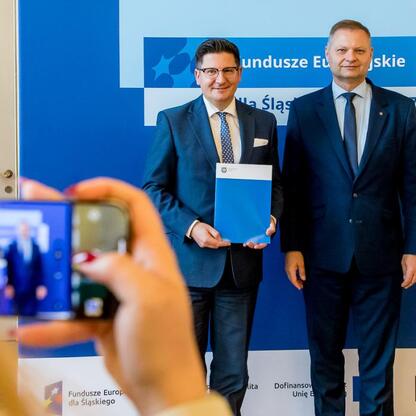 Członek Zarządu Województwa Śląskiego Rafał Adamczyk i inny mężczyzna w stroju formalnym prezentują dokumenty przed logo Funduszy Europejskich dla Śląska 2021-2027. (wygenerowano przez AI)