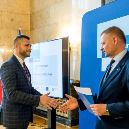 Dwóch członków Zarządu Województwa Śląskiego w strojach formalnych składają sobie uścisk dłoni, podczas ceremonii wręczenia listów gratulacyjnych. (wygenerowano przez AI)