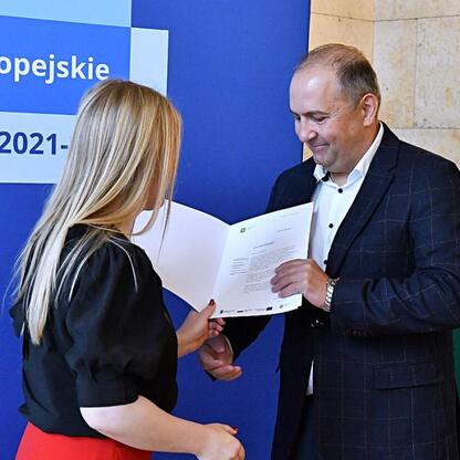 Dwie osoby w strojach formalnych wymieniają dokumenty, tło z napisem o funduszach europejskich dla Śląska 2021-2027. (wygenerowano przez AI)