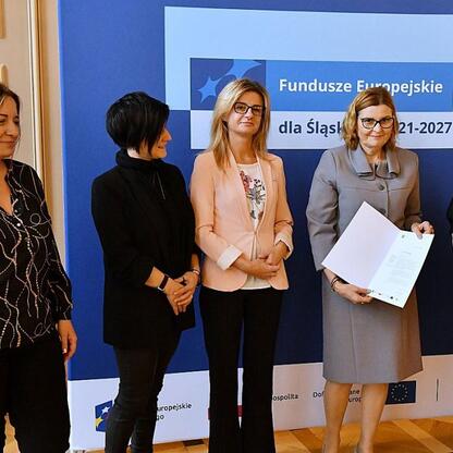 Pięciu członków Zarządu Województwa Śląskiego wręczają listy gratulacyjne beneficjentom projektów edukacyjnych finansowanych z funduszy europejskich. (wygenerowano przez AI)
