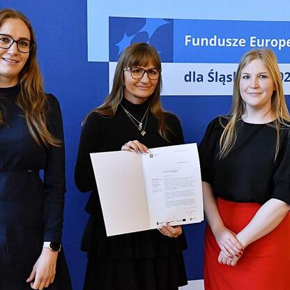 Trzy kobiety przedstawiają dokumenty, promując projekty finansowane przez Unią Europejską dla Śląska. (wygenerowano przez AI)