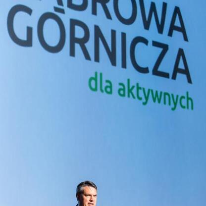 Na scenie stoi mówca w garniturze, trzymający mikrofon, na tle ekranu z napisem 