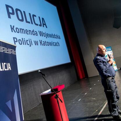 Na scenie stoi funkcjonariusz Policji, promujący bezpieczeństwo drogowe i pomoc w problemach młodych. (wygenerowano przez AI)