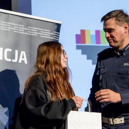 Na zdjęciu funkcjonariusz Policji i młoda kobieta podczas seminarium o bezpieczeństwie drogowym i cyberprzemocy w Dąbrowie Górniczej. (wygenerowano przez AI)