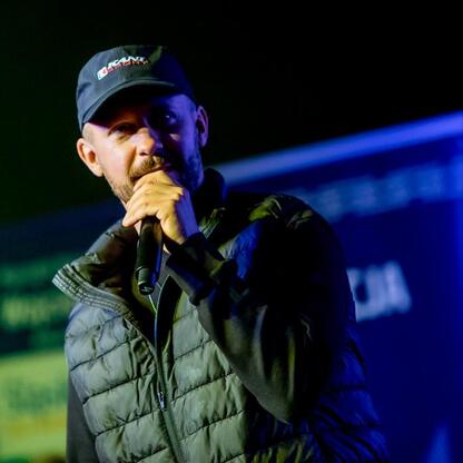 Na scenie w Dąbrowie Górniczej, podczas seminarium o bezpieczeństwie, mężczyzna w czapce i kurtce prezentuje się z mikrofonem, podkreślając temat bezpieczeństwa drogowego. (wygenerowano przez AI)
