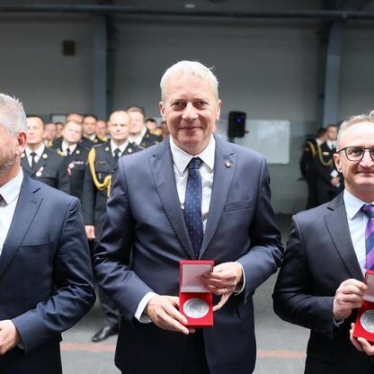 Trzech mężczyzn w strojach formalnych trzyma czerwone pudełka z medalami, uczestniczący w uroczystości honorującej strażaków. (wygenerowano przez AI)