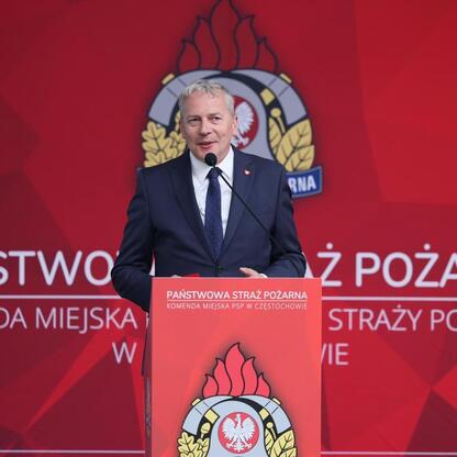 Marszałek Wojciech Saługa przemawia na podium w Komendzie Państwowej Straży Pożarnej w Częstochowie, omawiając inwestycje w regionie. (wygenerowano przez AI)