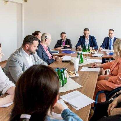Grupa osób w strojach formalnych siedzi wokół długiego drewnianego stołu, omawiając tematy związane z Radą Społeczną ds. Osób Niepełnosprawnych w Śląsku. (wygenerowano przez AI)
