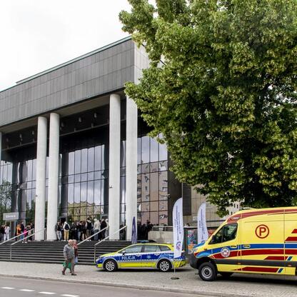 Modern building with banners, police car, ambulance, and gathered people, likely related to 