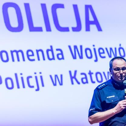 Policjant w mundurze przemawia na podium, trzymając mikrofon, z tłem 