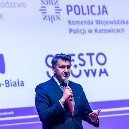 Na scenie mówca w ciemnym garniturze i czerwonym krawacie przemawia do publiczności, podkreślając bezpieczeństwo drogowe i problemy młodzieży. (wygenerowano przez AI)