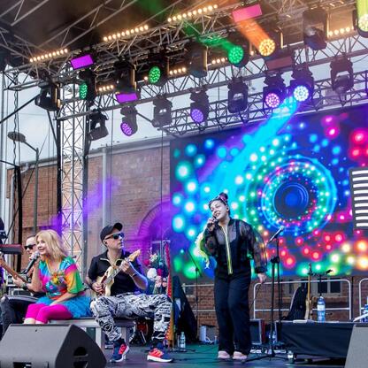 Zaprezentowany na zdjęciu jest koncert na festiwalu INDUSTRIADY, który uczcił przemysłowe dziedzictwo Śląska. Scena oświetlona kolorowymi światłami, muzykowie aktywnie grają, a publiczność cieszy się  (wygenerowano przez AI)