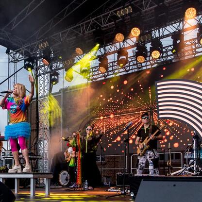 Widoczna jest scena koncertowa na festiwalu INDUSTRIADY, zielone drzewo i oświetlona sceną. (wygenerowano przez AI)