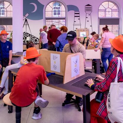Widoczna jest scena z festiwalu INDUSTRIADY, gdzie uczestnicy, w tym dzieci i dorosli, uczestniczą w interaktywnych aktywnościach przy stole, w otoczeniu kolorowych dekoracji i murali, w Fabryce Porce (wygenerowano przez AI)