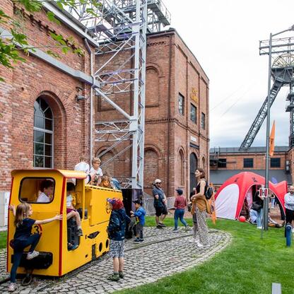 Widoczna jest scena festiwalowa w Carnall Sztolni Królowa Luiza w Zabrzu, podczas INDUSTRIADY 2023. W tle historyczne budynki przemysłowe, w pierwszym planie atrakcje interaktywne. (wygenerowano przez AI)