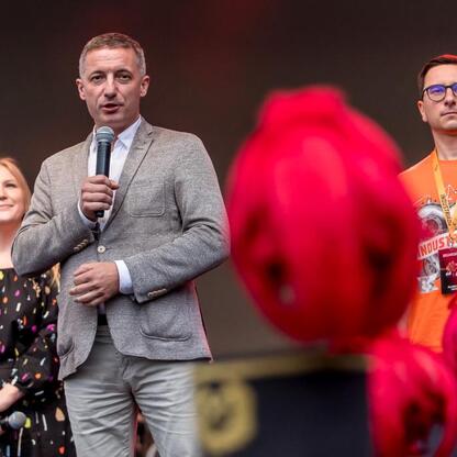 Na zdjęciu z 2023. edycji INDUSTRIADY, festiwalu upamiętniającego przemysłowe dziedzictwo Śląska, mężczyzna w szarym płaszczu przemawia do publiczności, podczas gdy drugi w pomarańczowej koszulce obse (wygenerowano przez AI)