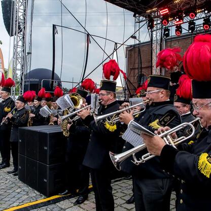 Zdjęcie przedstawia scenę z festiwalu INDUSTRIADY, odbywającego się w 2019 roku w Silesia. Na scenie występują muzycy w formacji, ubrani w czarne stroje z czerwonymi i złotymi akcentami, podczas konce (wygenerowano przez AI)