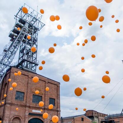 Widoczna jest scena z festiwalu INDUSTRIADY w Zabytkowej Kopalni Ignacy w Rybniku, gdzie balony w kolorach pomarańczowym i niebieskim wznoszą się w powietrze, symbolizując temat festiwalu 