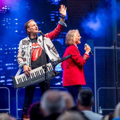 Zaprezentowane na zdjęciu moment z festiwalu INDUSTRIADY w Sosnowcu, gdzie dwaj artystowie w czerwonej i jeansowej kurtce wykonują muzykę na scenie. (wygenerowano przez AI)