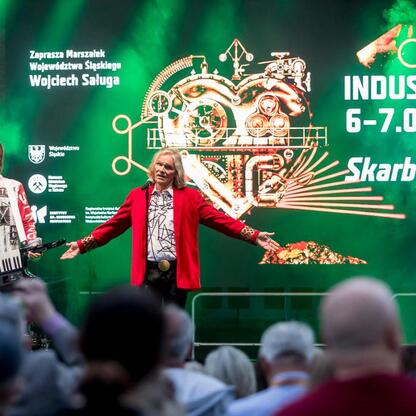 Fotografia przedstawia scenę z festiwalu INDUSTRIADA, z widocznymi wykonawcami i widownią, z napisem 
