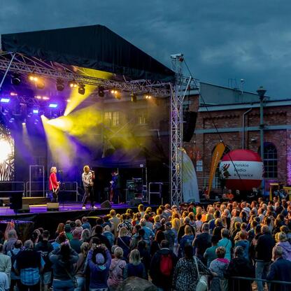 Widoczna jest scena koncertowa podczas festiwalu INDUSTRIADY, z kolorowymi oświetleniami i publicznością cieszącą się muzyką. (wygenerowano przez AI)