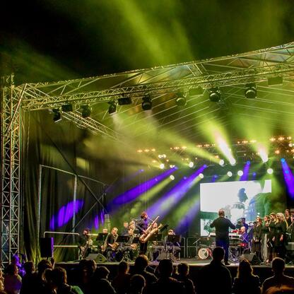 Fotografia przedstawia zakończenie festiwalu INDUSTRIADY w Zabytkowej Kopalni Ignacy w Rybniku. Scena oświetlona zielonymi i fioletowymi światłami, z muzykami i chórem. Publikacja cieszy się. (wygenerowano przez AI)