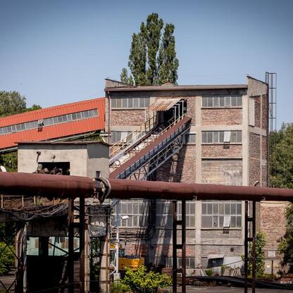 Zdjęcie przedstawia industrialny teren z czerwonym budynkiem i wielopoziomowym obiektem z wielkimi oknami, będący częścią dawnego kompleksu kopalni Wujek w Katowicach, Polska. (wygenerowano przez AI)