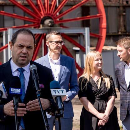 Grupa przedstawicieli z Polskiej Grupy Górniczej, Województwa Śląskiego, Katowic i GZM na konferencji prasowej przed kołem przemysłowym, omawiając przyszłość terenów po kopalni Wujek. (wygenerowano przez AI)