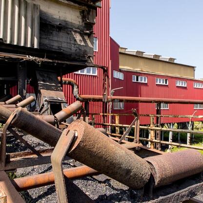 Abandoned industrial site, former coal mine, rusted pipes and machinery scattered, red and yellow corrugated metal buildings in the background, rural area with greenery surrounding the site. (wygenerowano przez AI)