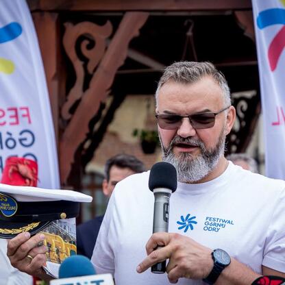 Na zdjęciu: Mężczyzna w białej koszulce z logo festiwalu, mówiący na konferencji prasowej w ramach 5. edycji Festiwalu Górnej Odry. (wygenerowano przez AI)