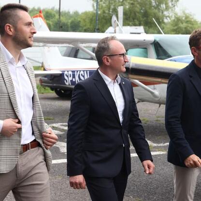 Trzech mężczyzn w garniturach spaceruje po tarmacze lotniska, omawiając Air Show w Rudnikach. (wygenerowano przez AI)
