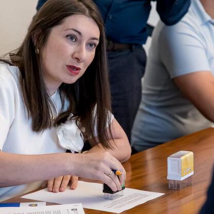 Kobieta w białej koszulce siedzi przy stole, wyjaśniając program 