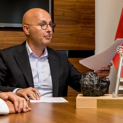 Mężczyzna w garniturze siedzi przy stole, trzymając dokument i wskazując na trofeum z węglem i motywami górniczymi. (wygenerowano przez AI)