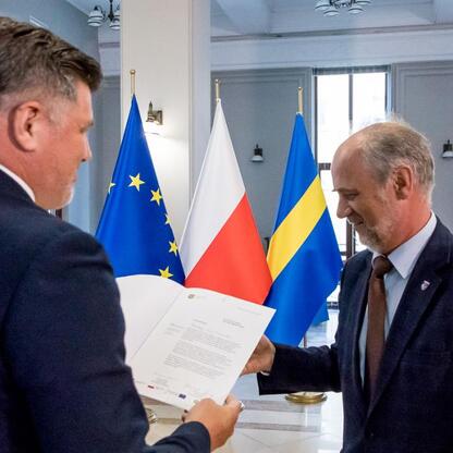 Dwa urzędnicy w strojach formalnych przekazują dokumenty w ceremonii na Śląsku, z flagami UE, Polski i Ukrainy. (wygenerowano przez AI)