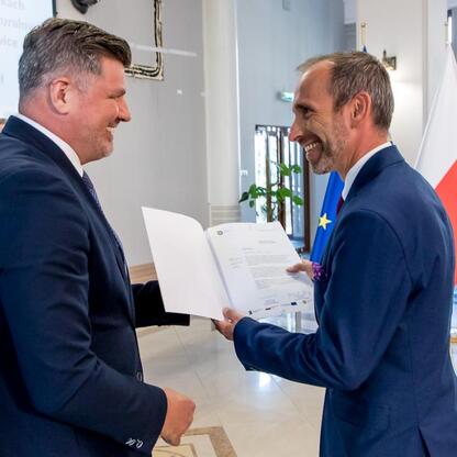 Dwa mężczyźni w strojach formalnych wymieniają dokumenty w budynku administracyjnym, zaznaczając sukces w projekcie energetycznym Śląska. (wygenerowano przez AI)
