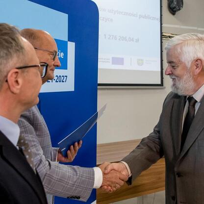 Trzech mężczyzn w strojach formalnych trzyma się za ręce przed logo i napisami programu Funduszy Europejskich dla Śląska 2021-2027. (wygenerowano przez AI)