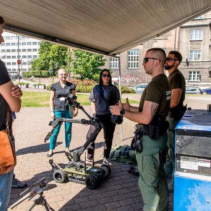 Grupa osób w mundurach i cywilnych ubraniach zgromadzona pod namiotem w mieście, uczestnicząc w ćwiczeniach kryzysowych. (wygenerowano przez AI)