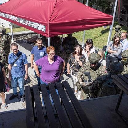 Grupa osób w mundurach i z sprzętem wojskowym na tle czerwonej namiotu z napisem 